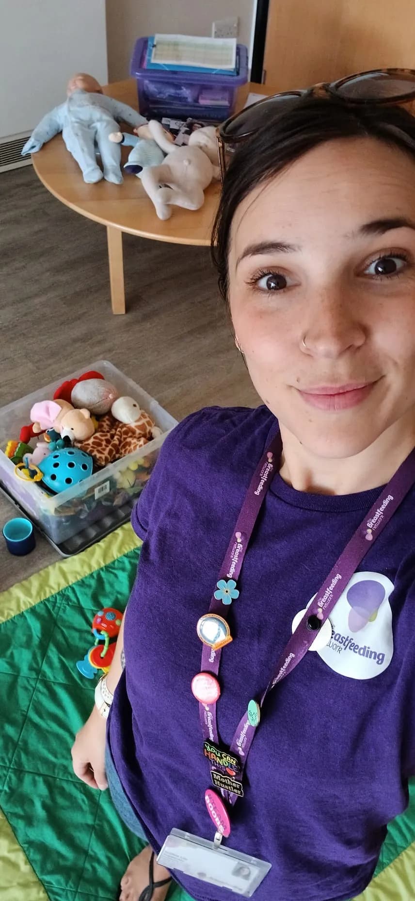 A woman in a purple shirt wearing a Breastfeeding lanyard smiles at the camera, with baby dolls and toys visible in the background.