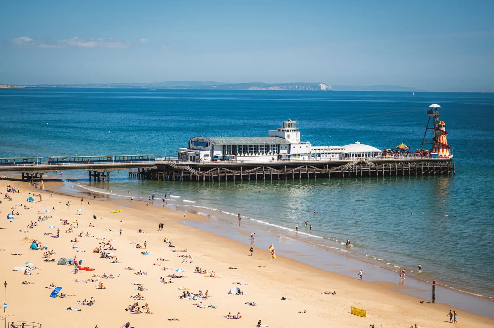 A long pier with buildings extends over blue ocean water from a crowded sandy beach under a clear sky.